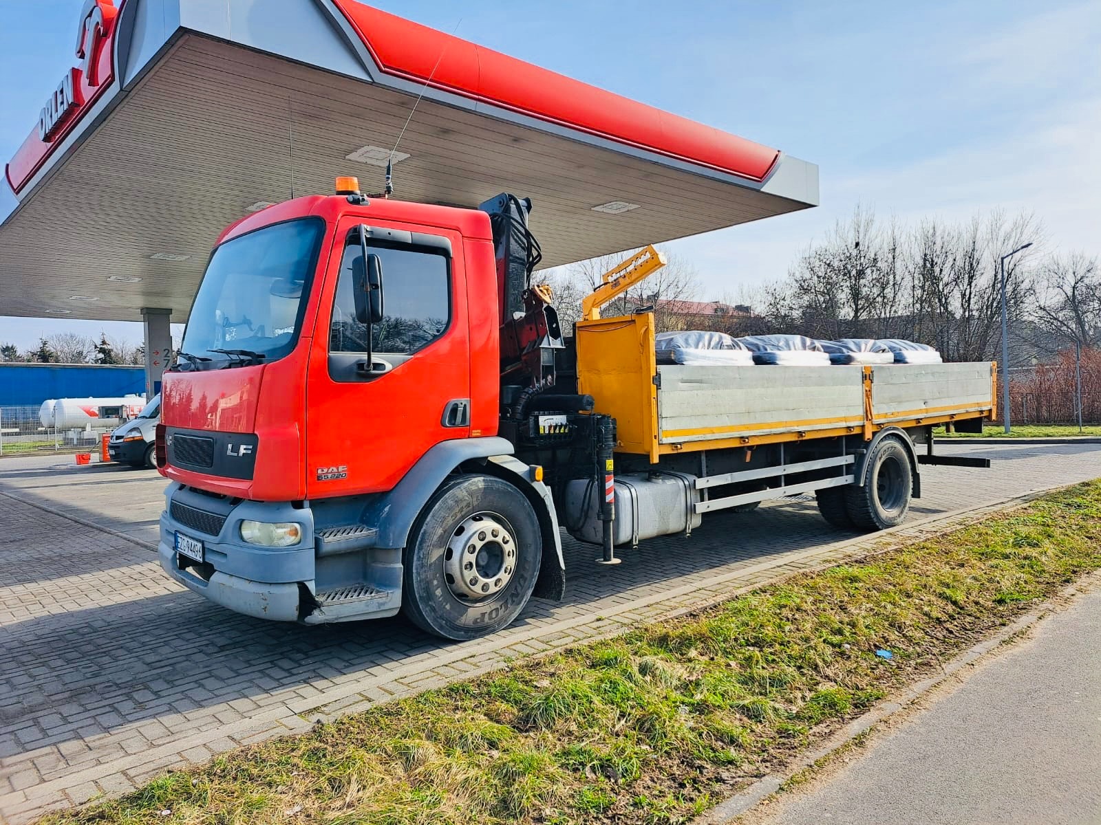 Czerwona ciężarówka DAF LF z żółtym dźwigiem i ładunkiem na pace, stoi na stacji benzynowej w słoneczny dzień. Widoczne detale: opona Dunlop, tablica rejestracyjna.