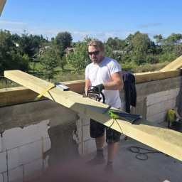 GibDach - Cieśla w okularach tnie deskę piłą łańcuchową na budowie więźby dachowej. W tle zielona roślinność i błękitne niebo. Widoczne narzędzia i elementy konstrukcyjne.