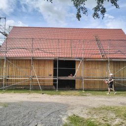 GibDach - Budynek w trakcie krycia czerwoną dachówką, otoczony rusztowaniami. Widoczny pracownik z narzędziem. Częściowo drewniana konstrukcja, błękitne niebo w tle.