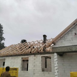 GibDach - Budowa domu z białych bloczków: widoczna drewniana więźba dachowa, komin i materiały budowlane Ytong. Etap wznoszenia konstrukcji, pochmurne niebo w tle.