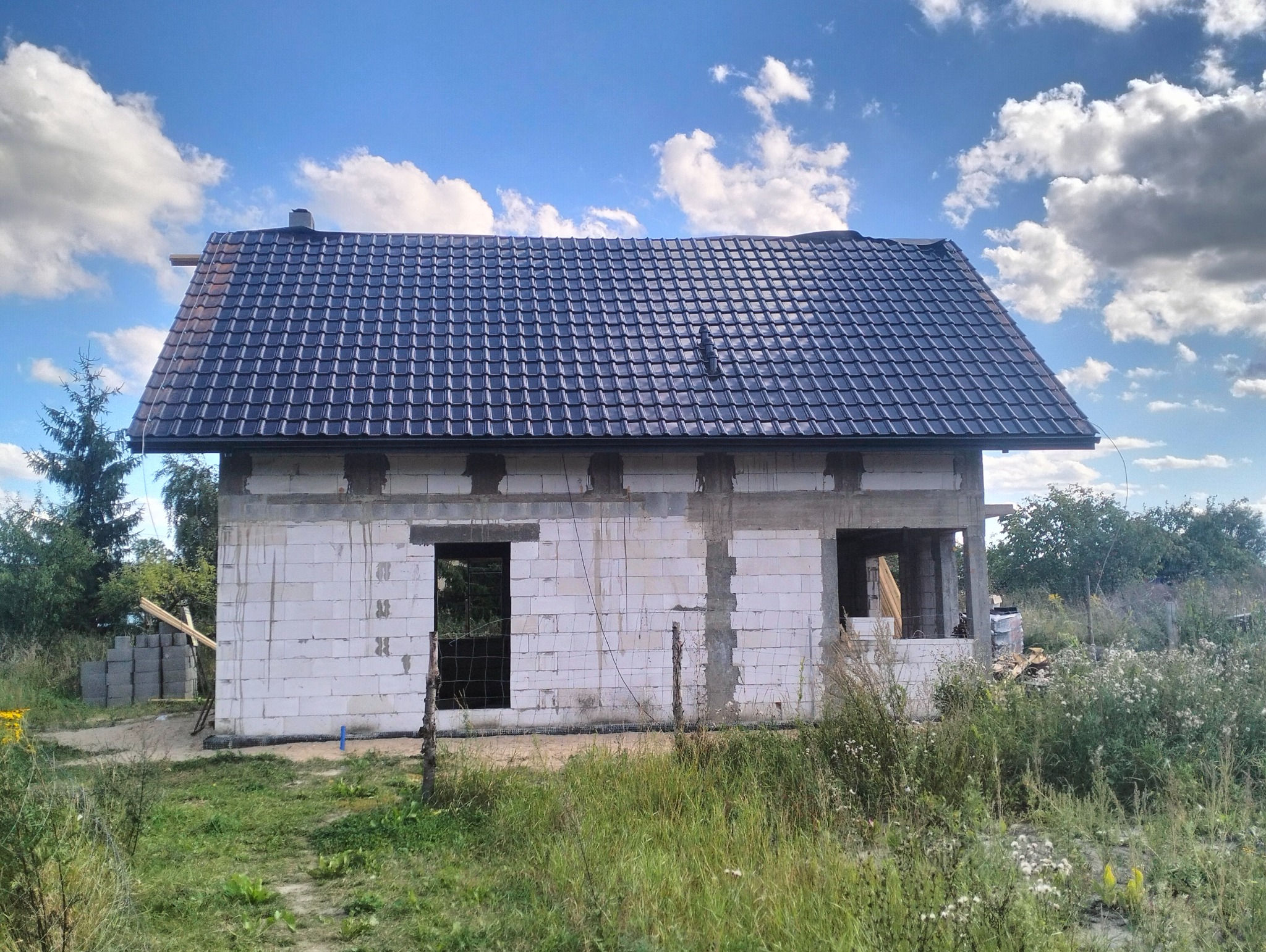 Dom w trakcie budowy z ciemną dachówką, widoczne ściany z bloczków i niezadaszona przestrzeń. Elementy konstrukcyjne dachu wystają poza obrys. Błękitne niebo w tle.