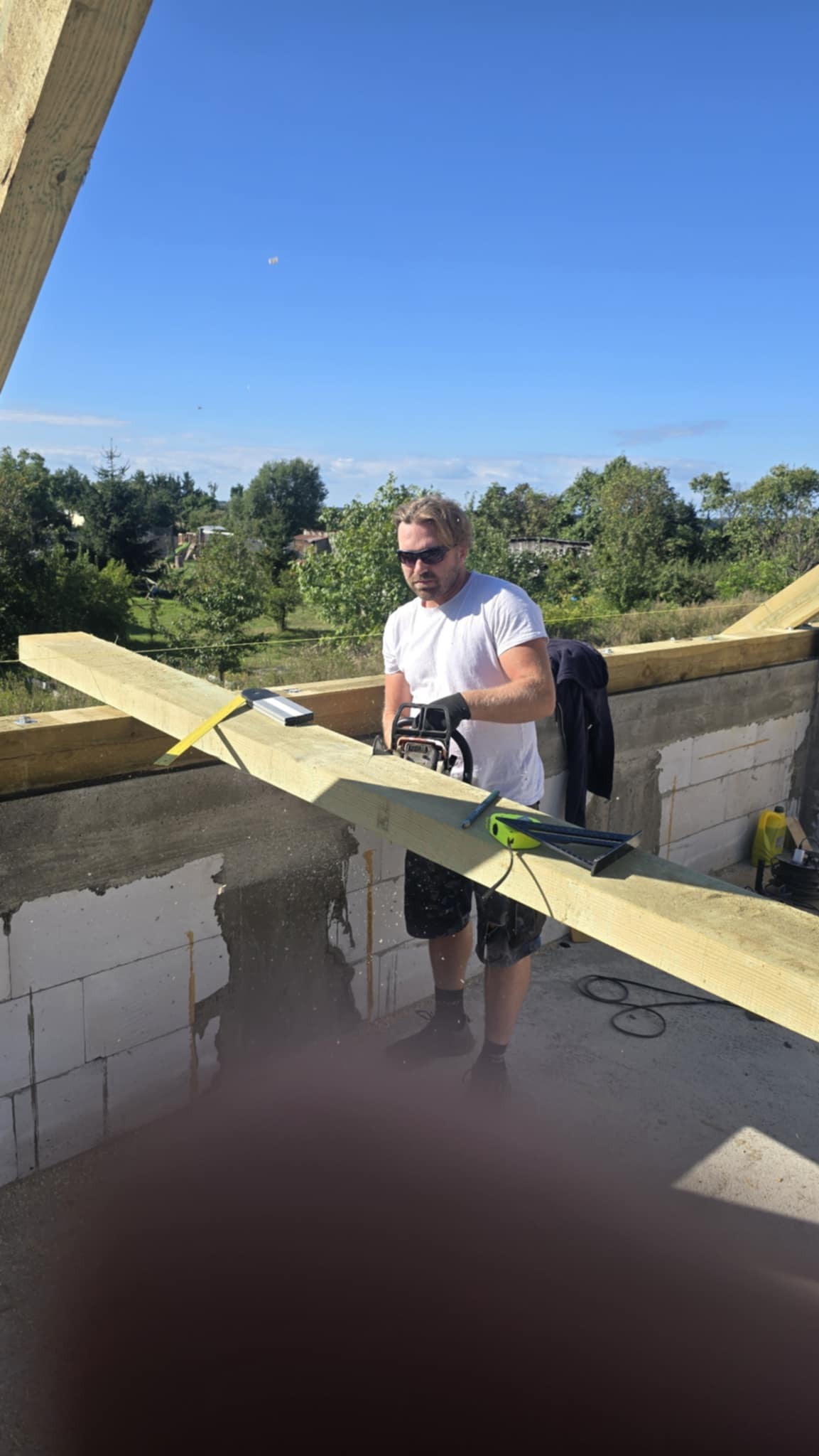 Cieśla w okularach tnie deskę piłą łańcuchową na budowie więźby dachowej. W tle zielona roślinność i błękitne niebo. Widoczne narzędzia i elementy konstrukcyjne.