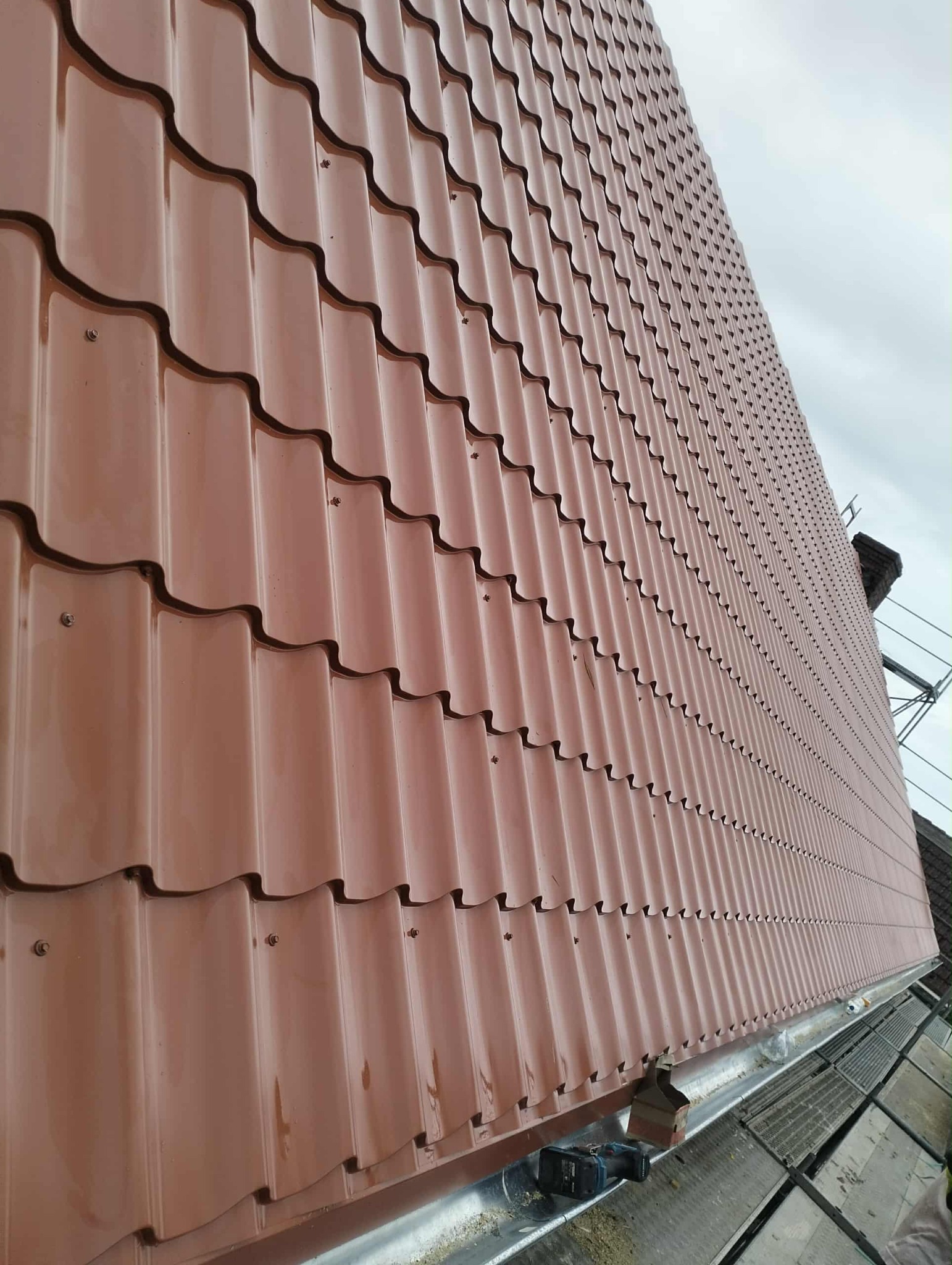 Nowe, brązowe dachówki ułożone na dachu, widoczne z dołu pod kątem. Na pierwszym planie rynna i narzędzia. W tle rusztowanie i fragment sąsiedniego budynku.