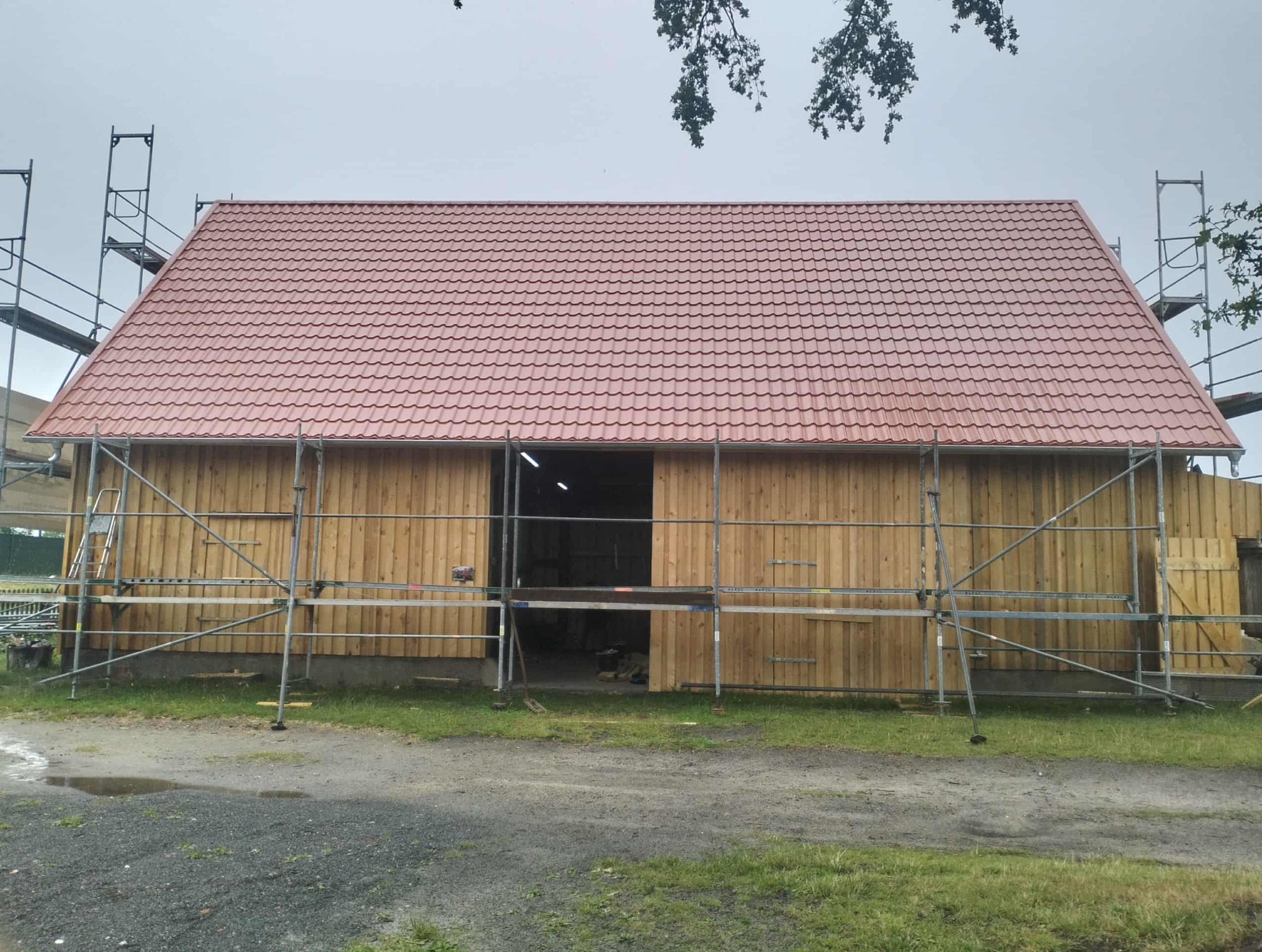 Budynek z nowym, czerwonym dachem z dachówki, otoczony rusztowaniami. Widoczne drewniane ściany i fragment zieleni. Pochmurne niebo w tle.