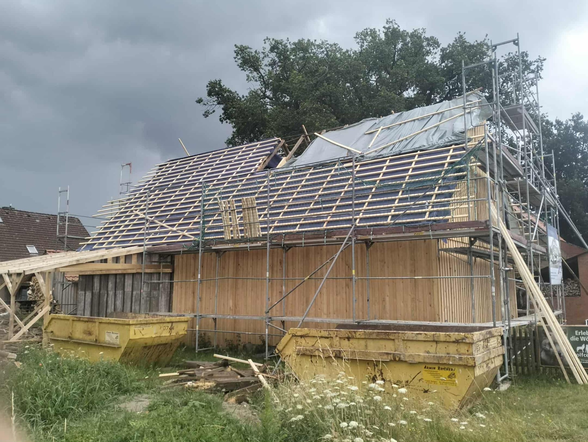 Budowa dachu z widoczną konstrukcją drewnianą i rusztowaniami. Dwa żółte kontenery na gruz na pierwszym planie. Pochmurne niebo w tle.