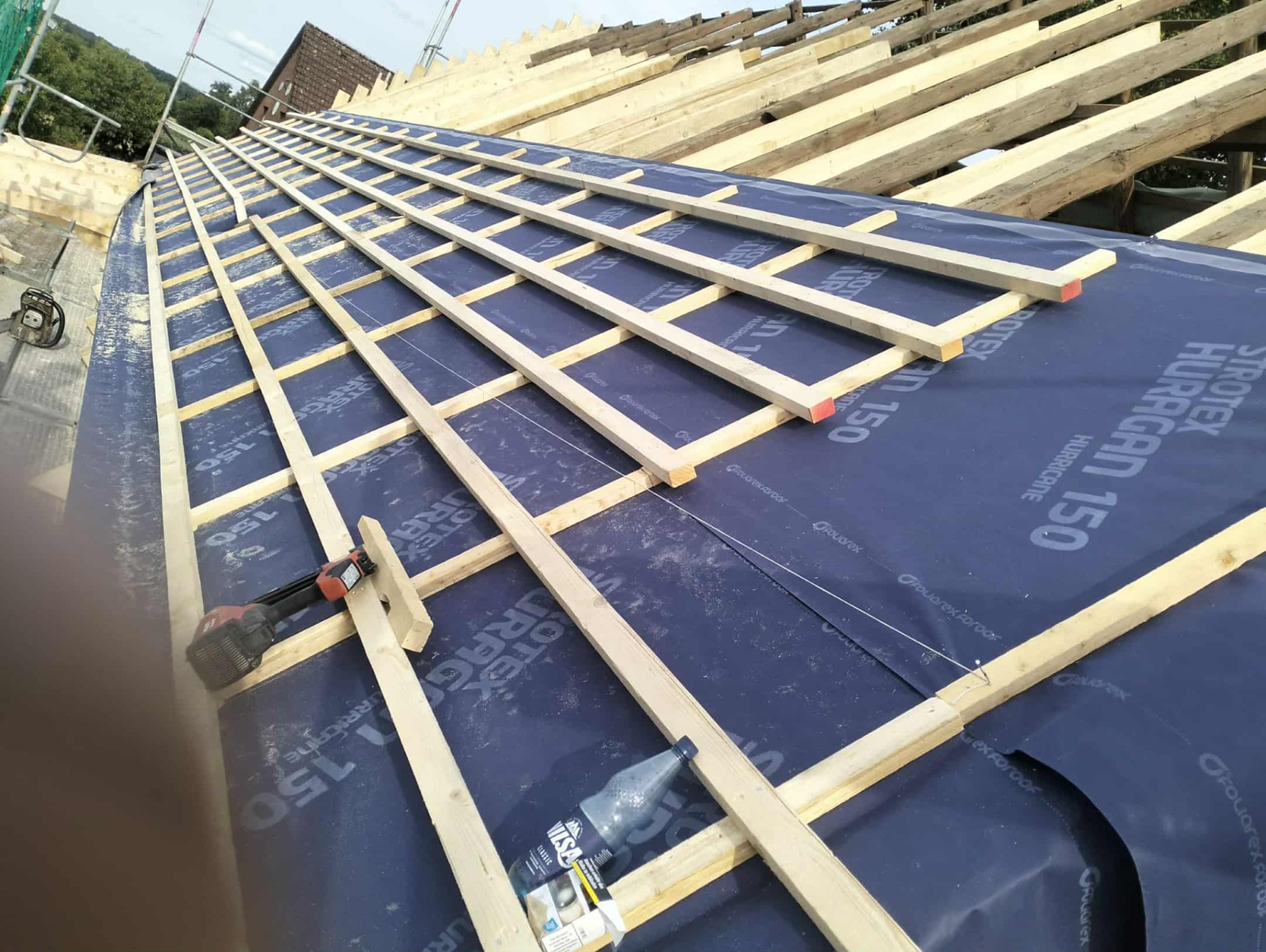Konstrukcja dachu w trakcie budowy: drewniane łaty na membranie Strotex Huragan 150, narzędzia i napój w tle. Widok z góry na budowę dachu.