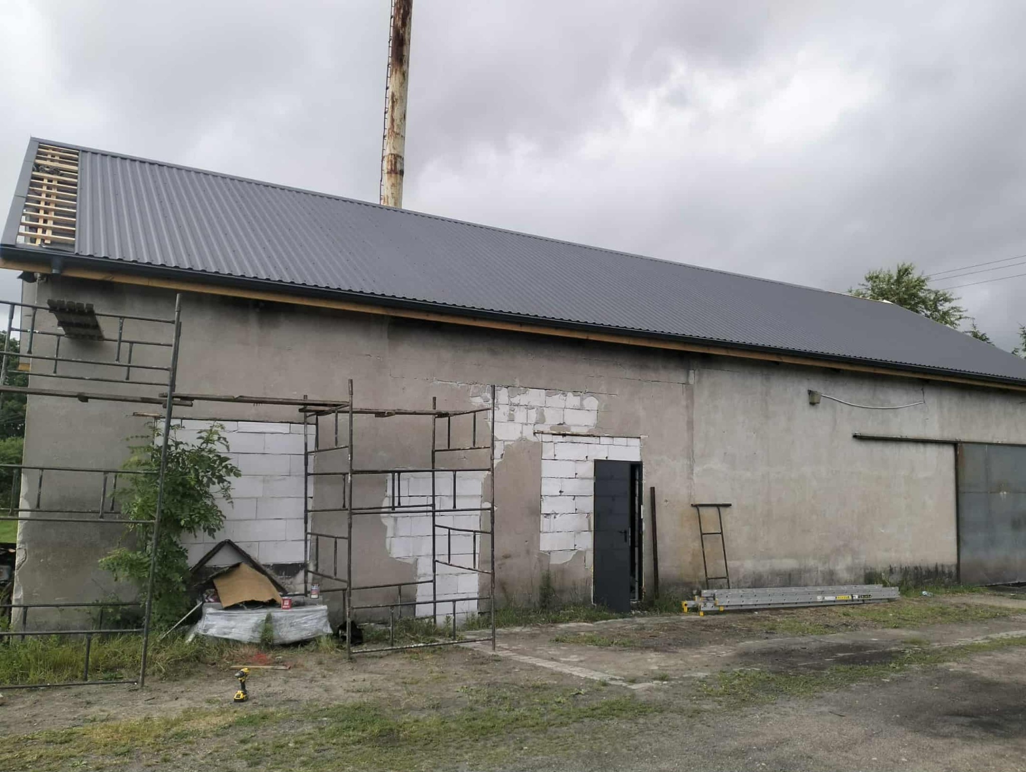 Szary dach z blachy trapezowej na budynku gospodarczym, częściowo z drewnianą konstrukcją, widoczne rusztowania i narzędzia, pochmurne niebo w tle.