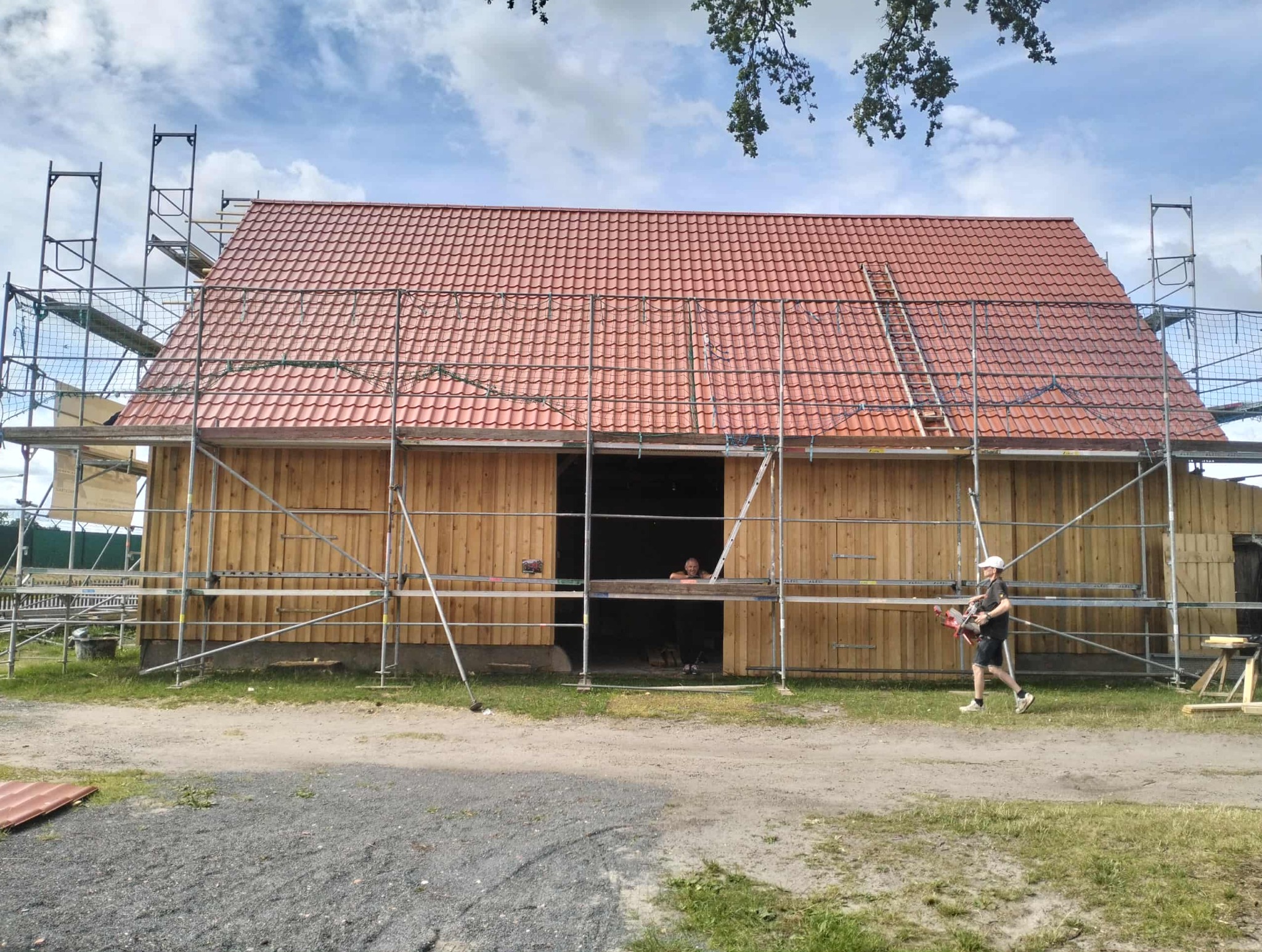 Budynek w trakcie krycia czerwoną dachówką, otoczony rusztowaniami. Widoczny pracownik z narzędziem. Częściowo drewniana konstrukcja, błękitne niebo w tle.
