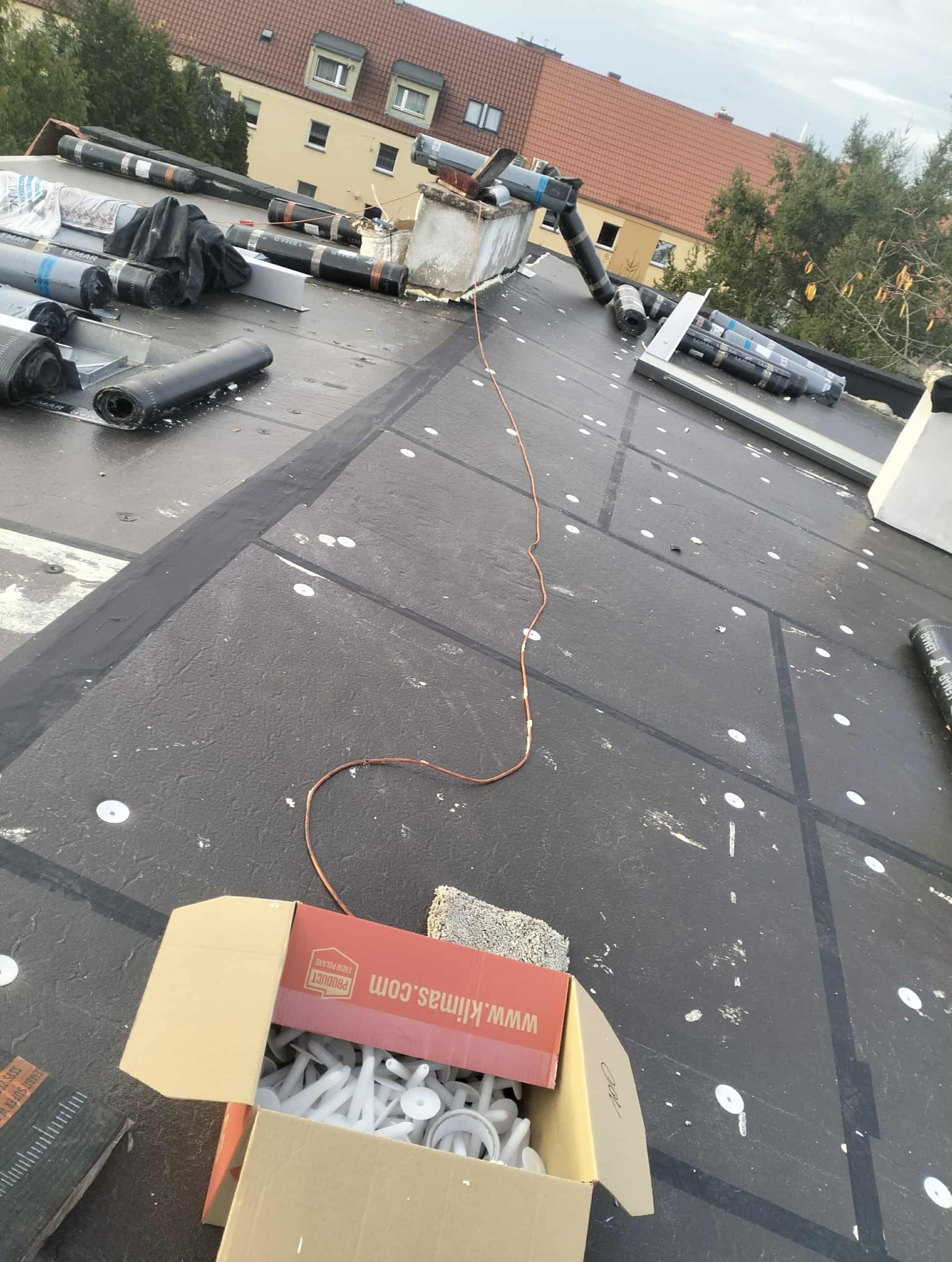 Czarny, płaski dach w trakcie prac. Widoczne rolki papy, elementy wentylacji, przewód uziemiający i karton z łącznikami. W tle budynek mieszkalny.