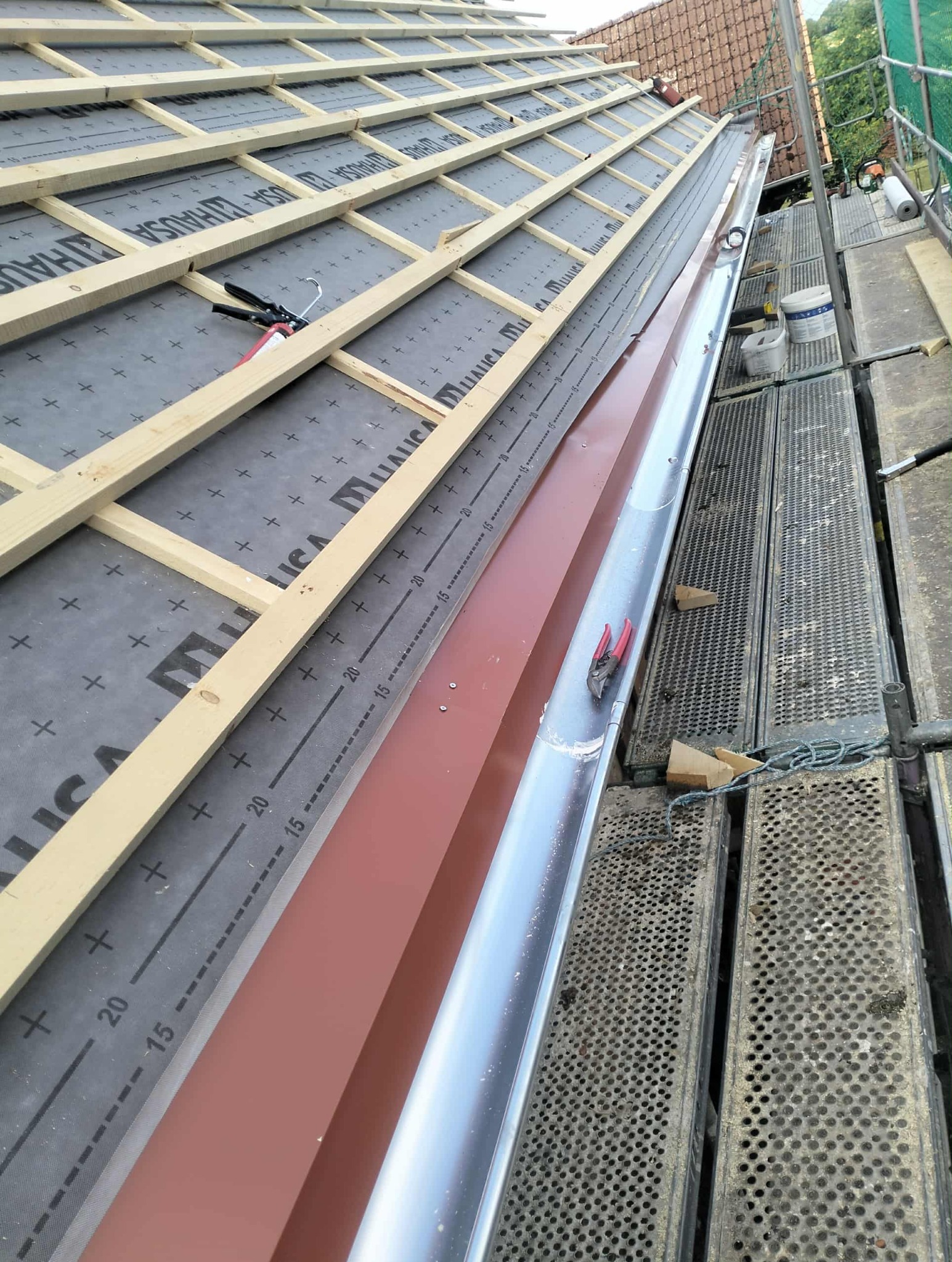 Montaż rynny na dachu z membraną i łatami. Widoczne narzędzia dekarskie i rusztowanie. Prace dekarskie w toku, z bliska, z perspektywy wykonawcy.