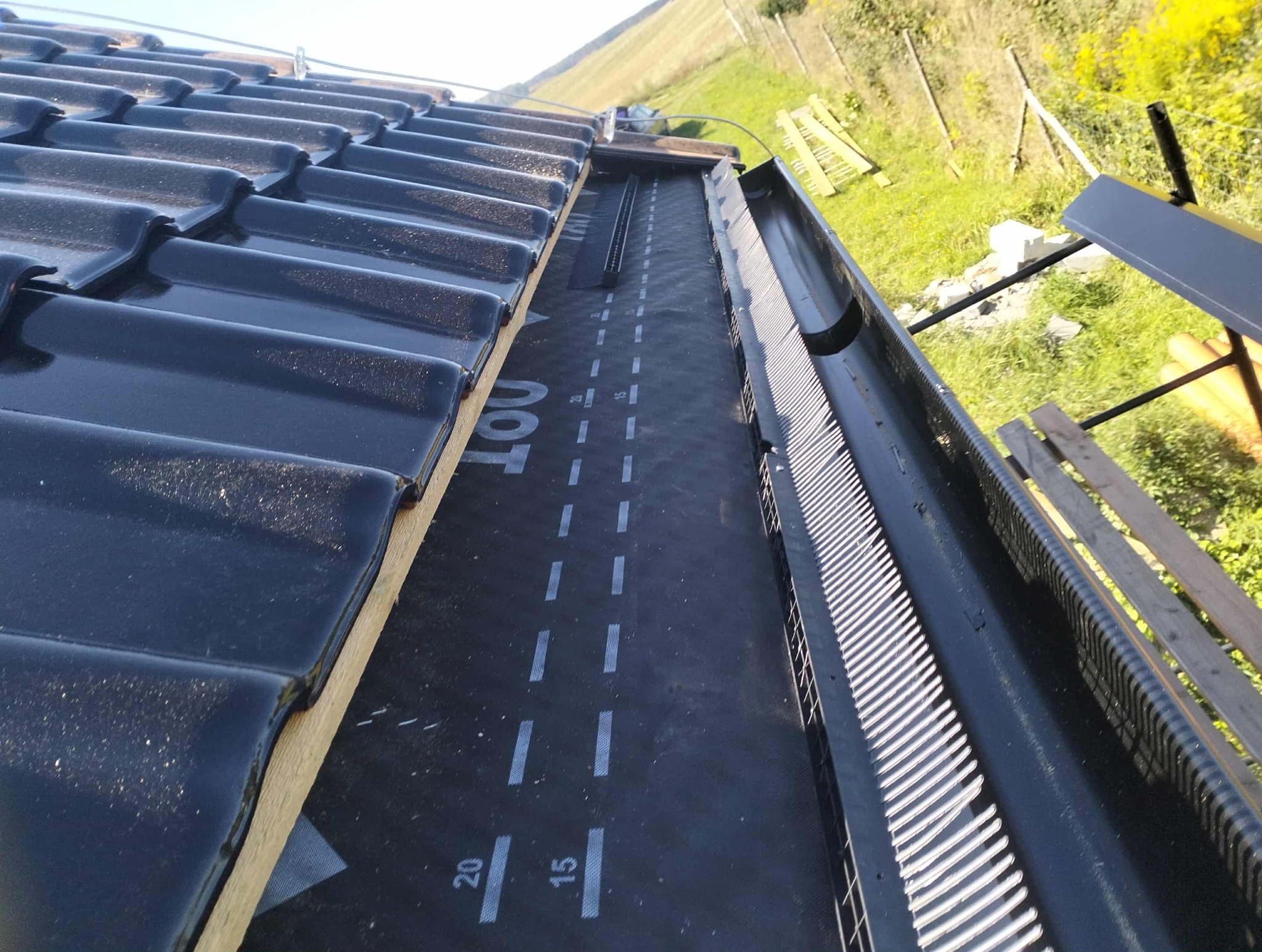 Czarny dach z połyskującą dachówką, widoczna membrana dachowa z oznaczeniami i zamontowana rynna z zabezpieczeniem przed liśćmi. Ujęcie z góry, w tle zieleń.