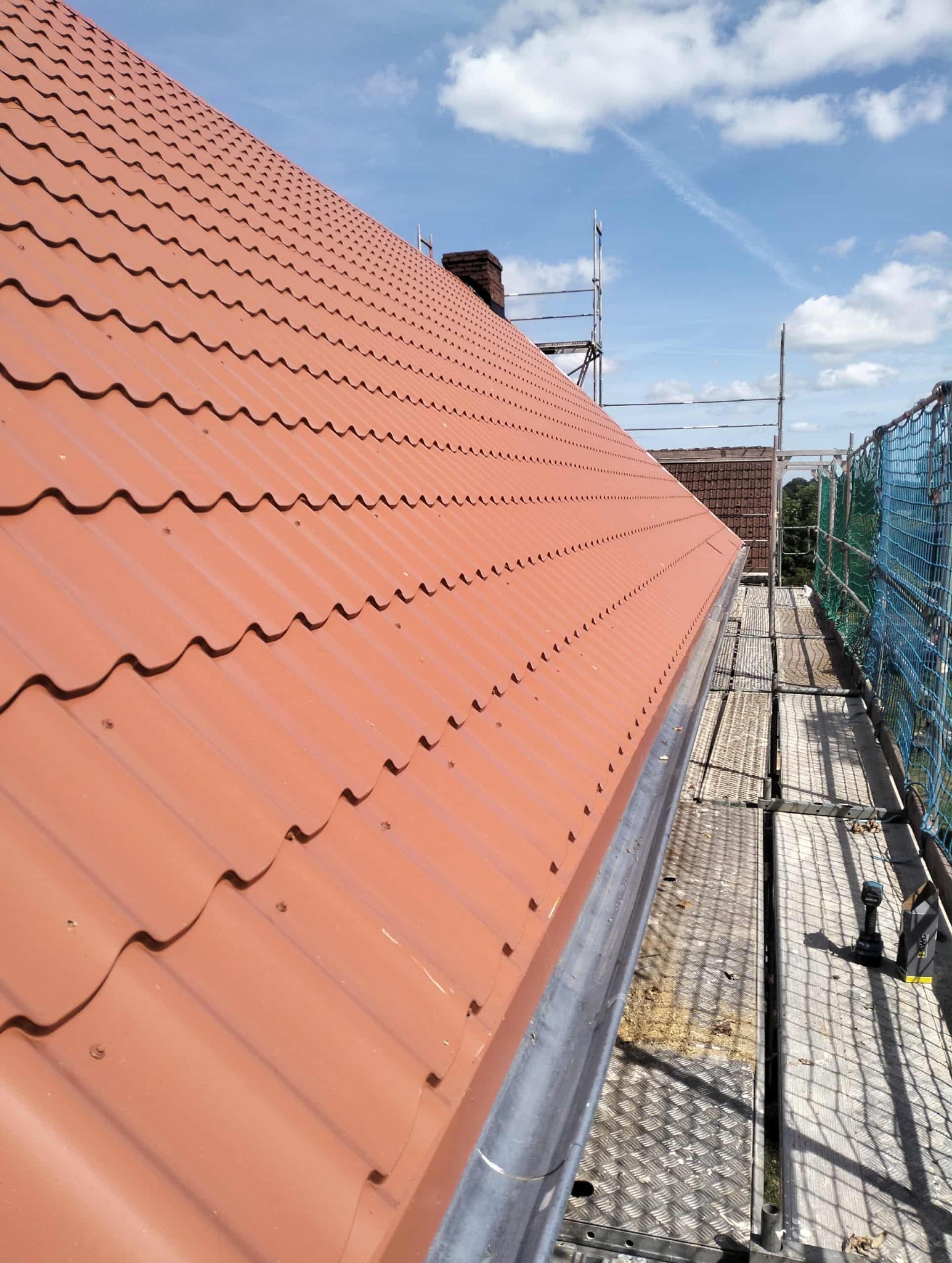 Czerwona dachówka ceramiczna na skośnym dachu, widok z góry, z zamontowaną rynną i rusztowaniem w tle, przy słonecznej pogodzie.