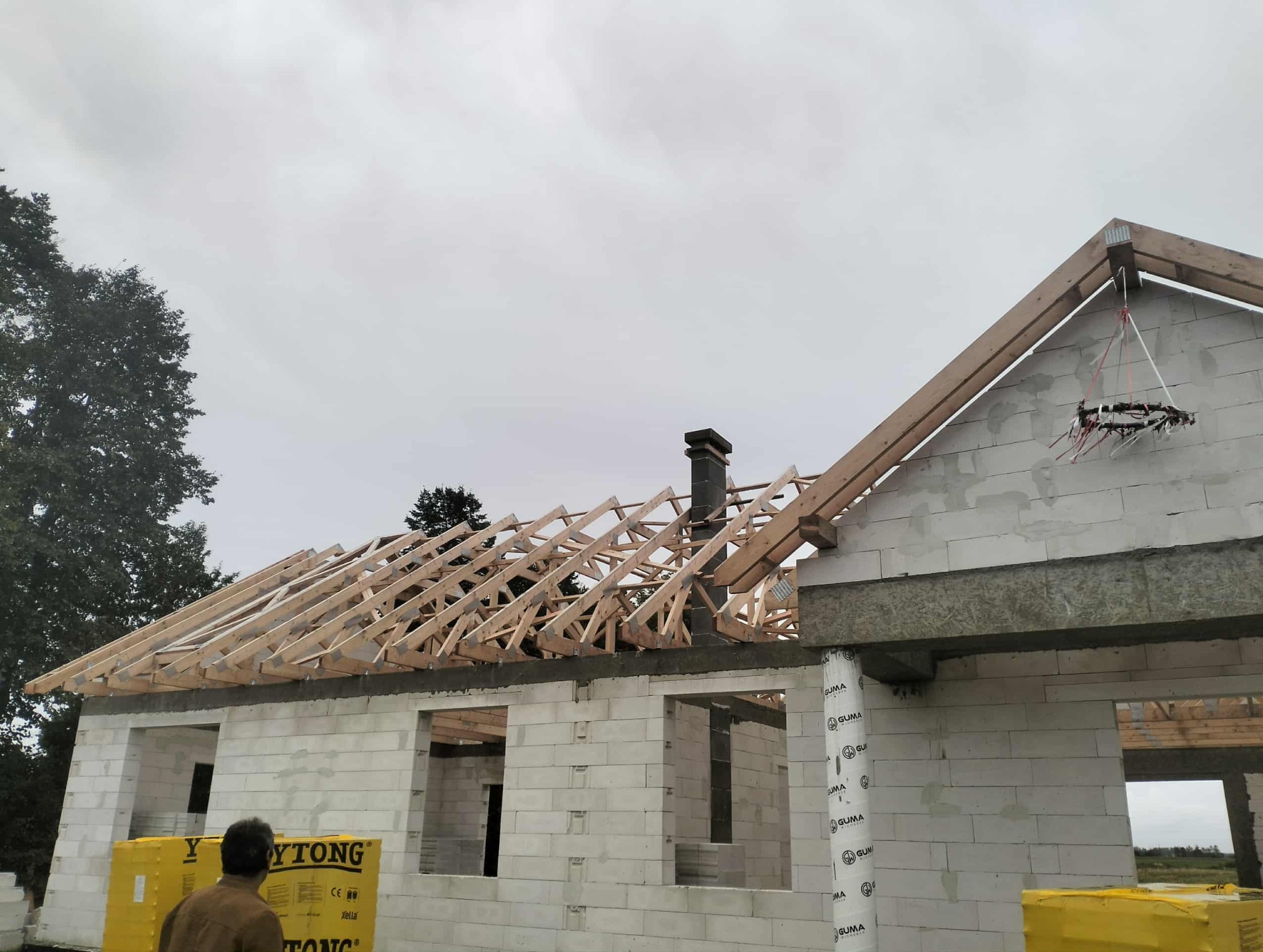 Budowa domu z białych bloczków: widoczna drewniana więźba dachowa, komin i materiały budowlane Ytong. Etap wznoszenia konstrukcji, pochmurne niebo w tle.