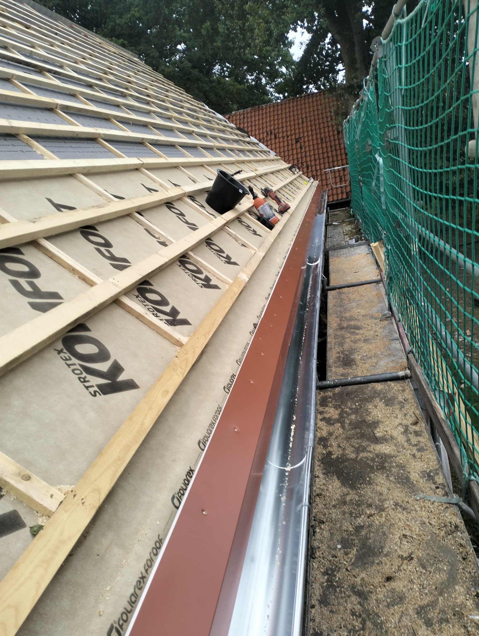 Dach w trakcie budowy: widoczna membrana, łaty, kontrłaty, narzędzia i fragment rynny. Ujęcie z góry, z boku, z widoczną siatką zabezpieczającą.