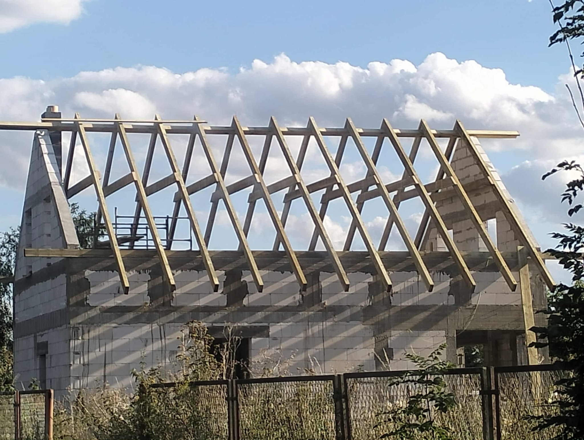 Wznoszenie więźby dachowej z drewnianych krokwi na murowanym budynku w stanie surowym otwartym, widok zza ogrodzenia, błękitne niebo w tle.