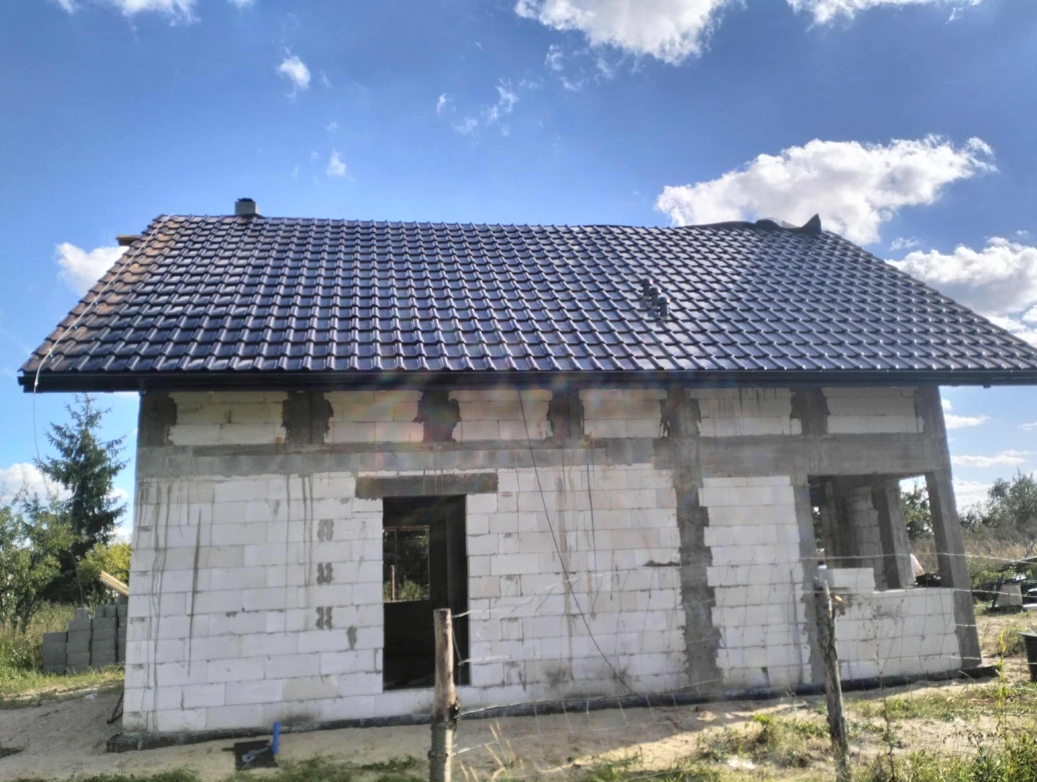 Dom w trakcie budowy z ciemną dachówką, widoczne niezadaszone okna i drzwi. Budynek z białych bloczków, na tle błękitnego nieba z chmurami.