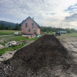 KOPCAR - Utwardzona droga do domu w trakcie budowy. Widoczny gruz, ziemia i traktor. W tle góry i pochmurne niebo. Prace ziemne w toku.