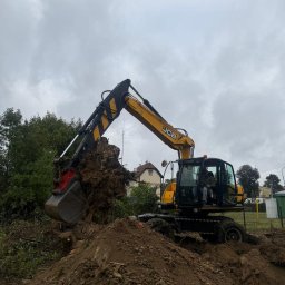 KOPCAR - Żółto-czarna koparka JCB z operatorem, unosi wykopany korzeń drzewa na tle pochmurnego nieba i zabudowań. Wokół hałda ziemi.