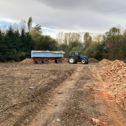 KOPCAR - Niebieski traktor z przyczepą na placu budowy, z gruzem i ziemią. Widoczne ślady opon i przygotowany teren pod dalsze prace. W tle drzewa i pochmurne niebo.