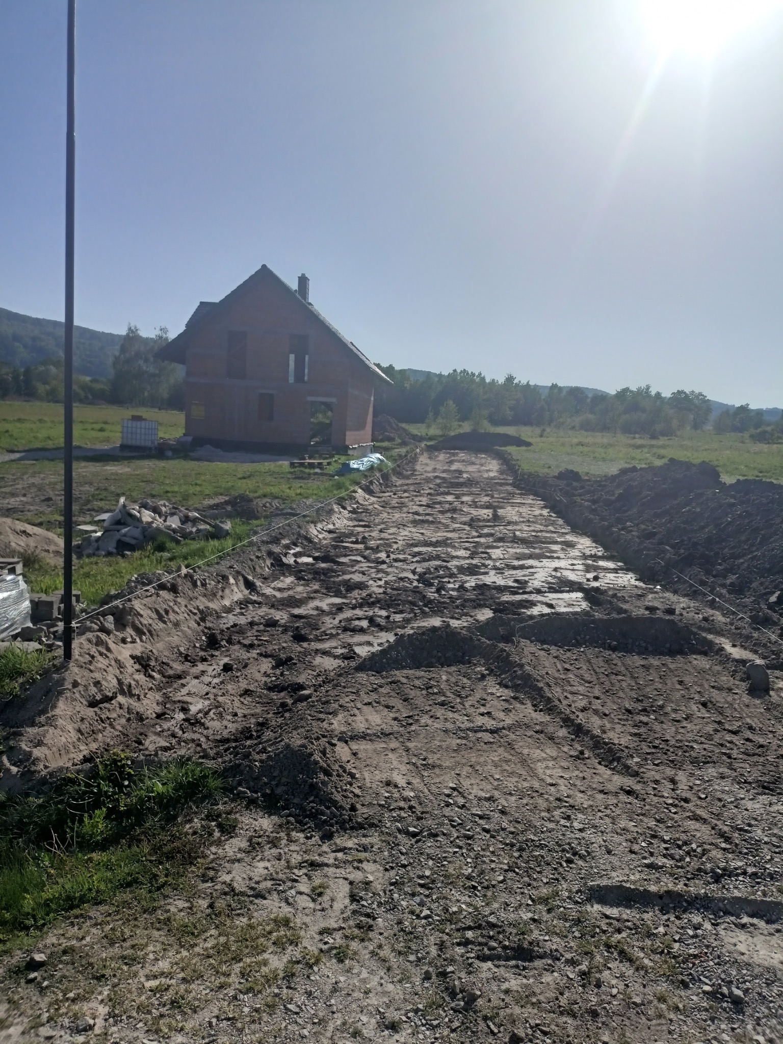 Przygotowanie terenu pod budowę domu: wytyczona i częściowo utwardzona droga dojazdowa, widoczny dom w stanie surowym otwartym, pagórkowate tereny w tle.
