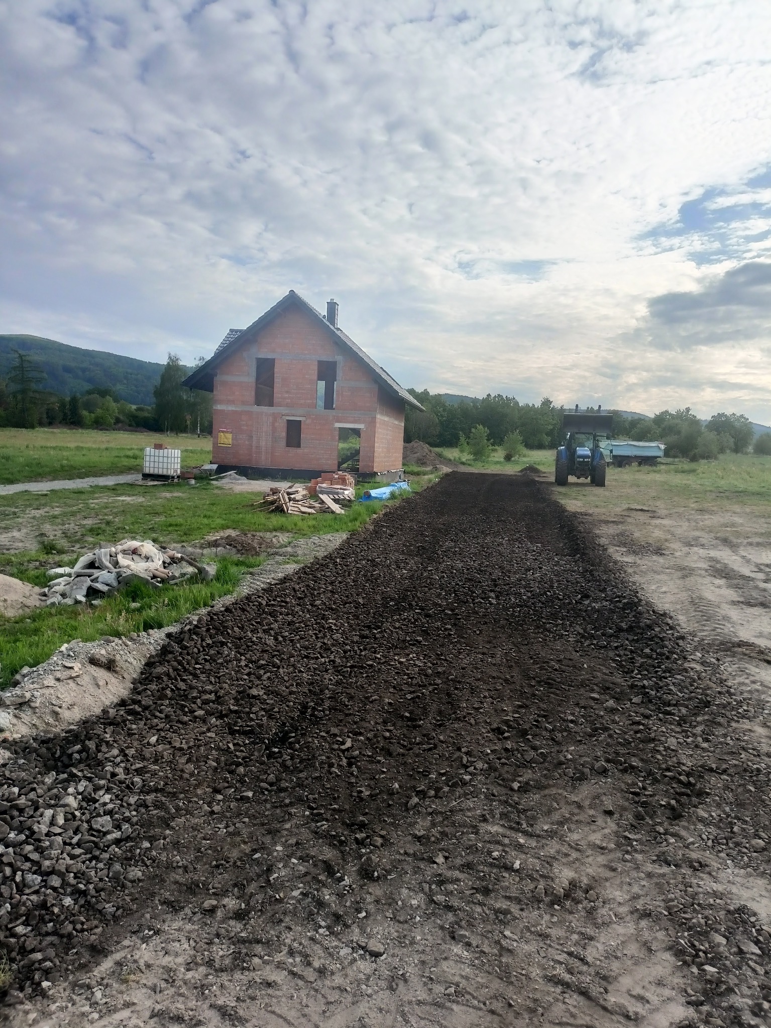 Utwardzona droga do domu w trakcie budowy. Widoczny gruz, ziemia i traktor. W tle góry i pochmurne niebo. Prace ziemne w toku.