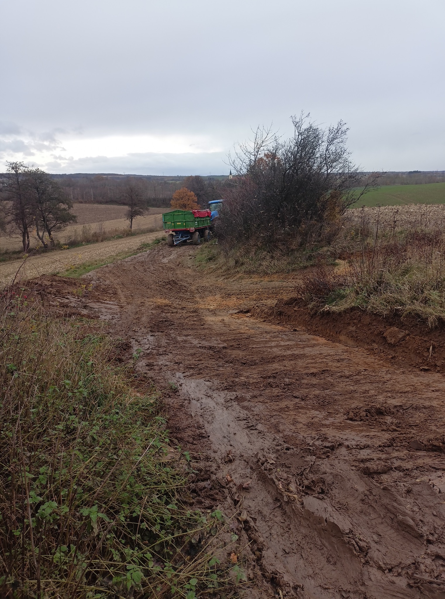 Błotnista droga gruntowa z koleinami, w tle traktor z przyczepą na polu pod pochmurnym niebem. Widoczne ślady opon i świeżo naruszona ziemia.
