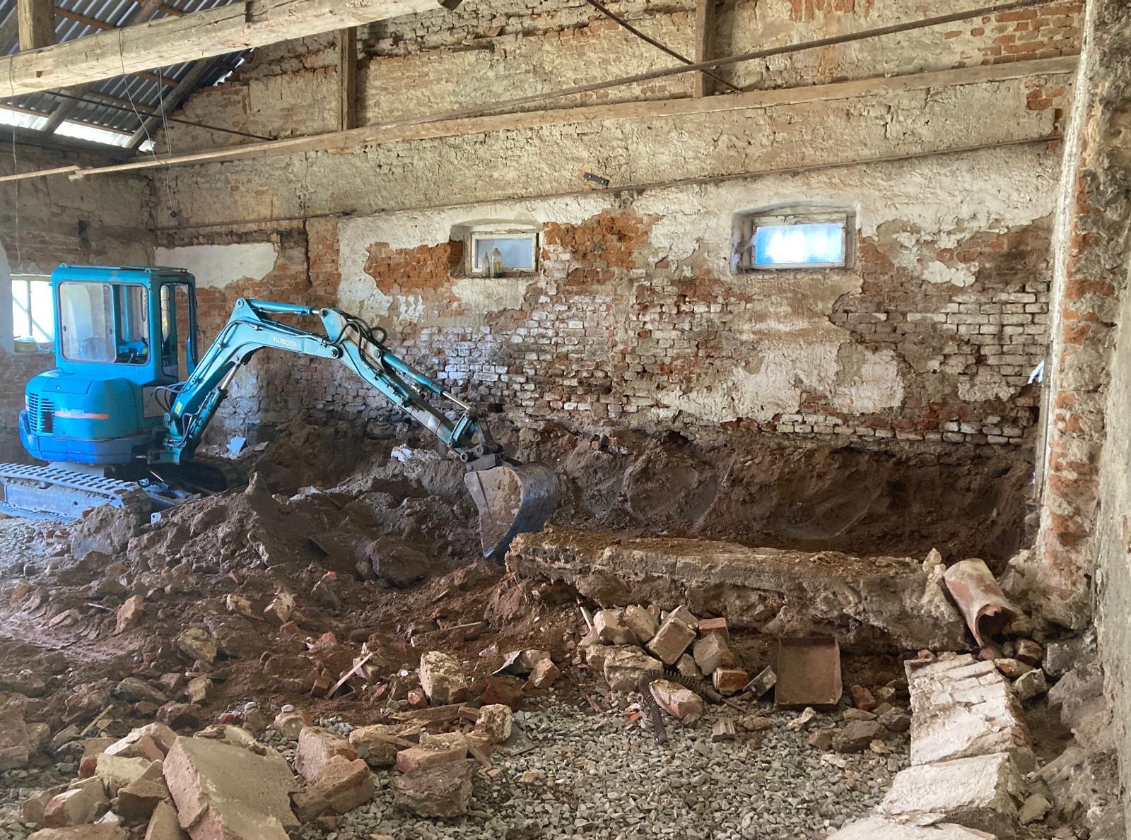 Niebieska mini koparka Kubota wewnątrz starego budynku z cegły, wykonująca wykop pod fundamenty. Widoczne okna i gruz.