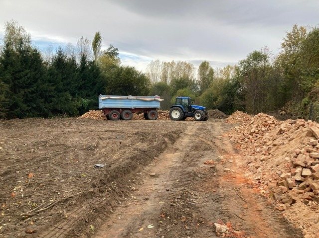 Niebieski traktor z przyczepą na placu budowy, z gruzem i ziemią. Widoczne ślady opon i przygotowany teren pod dalsze prace. W tle drzewa i pochmurne niebo.