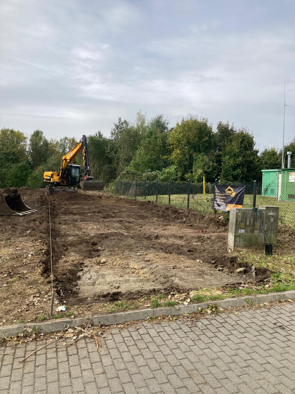 Żółta koparka JCB na działce po niwelacji terenu, widoczny wykop pod fundamenty. W tle zieleń drzew i krzewów, ogrodzenie z siatki i budynek techniczny.