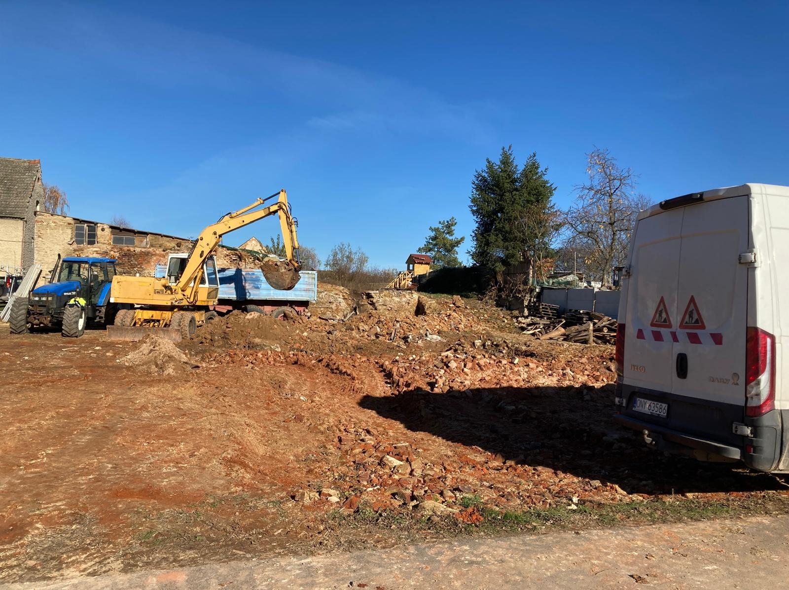 Żółta koparka ładuje gruz do niebieskiej ciężarówki na placu budowy. W tle budynek, drzewa i biały bus. Błękitne niebo.