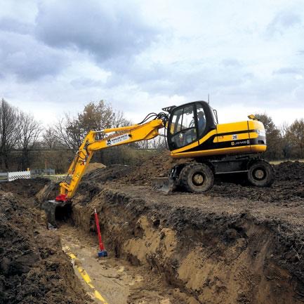 Żółta koparka kołowa w trakcie prac ziemnych, widoczny wykop pod rurę. Maszyna na tle pochmurnego nieba i drzew.