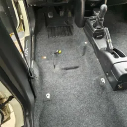 Szara, wyprana wykładzina podłogowa w samochodzie Nissan Note. Widoczne elementy wnętrza, pedały, dźwignia zmiany biegów. Czysta, odświeżona powierzchnia.