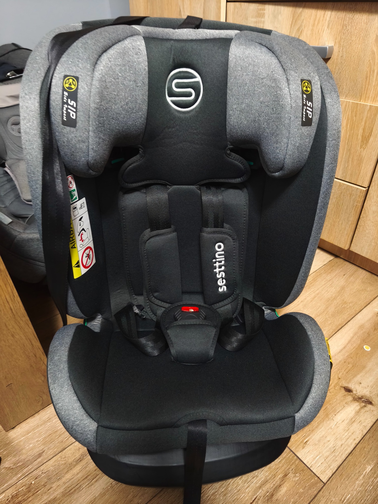 Szaro-czarny fotelik samochodowy Sesttino z widocznymi oznaczeniami bezpieczeństwa, umieszczony na drewnianej podłodze, gotowy do użytku.