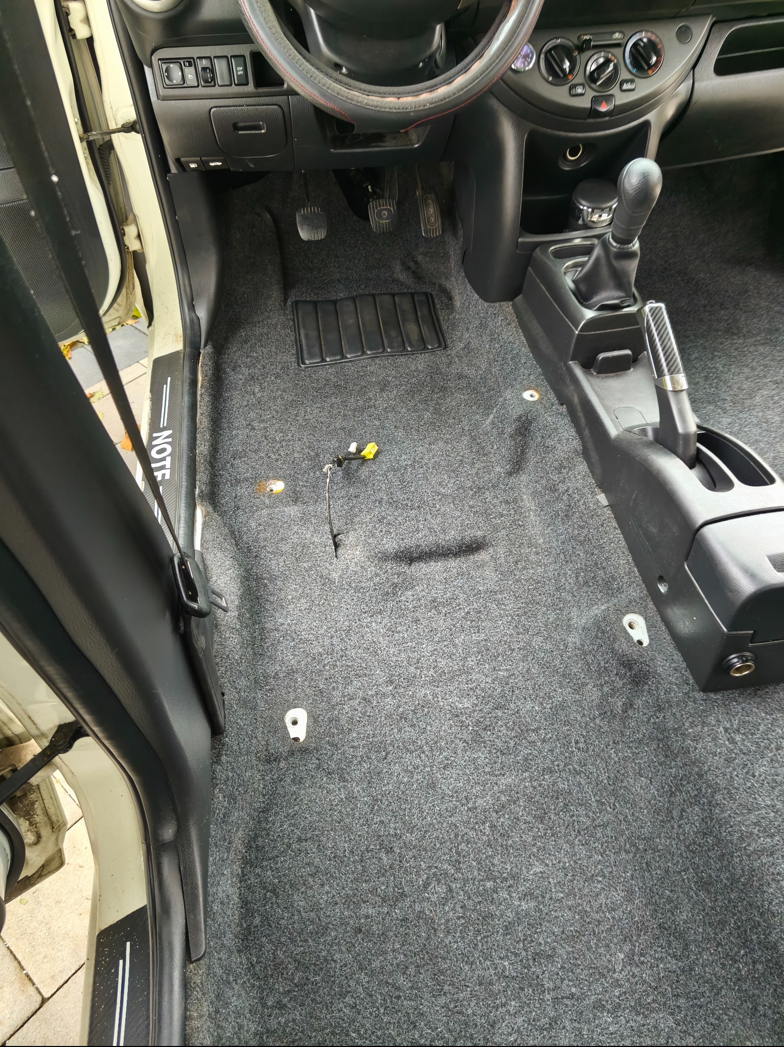 Szara, wyprana wykładzina podłogowa w samochodzie Nissan Note. Widoczne elementy wnętrza, pedały, dźwignia zmiany biegów. Czysta, odświeżona powierzchnia.