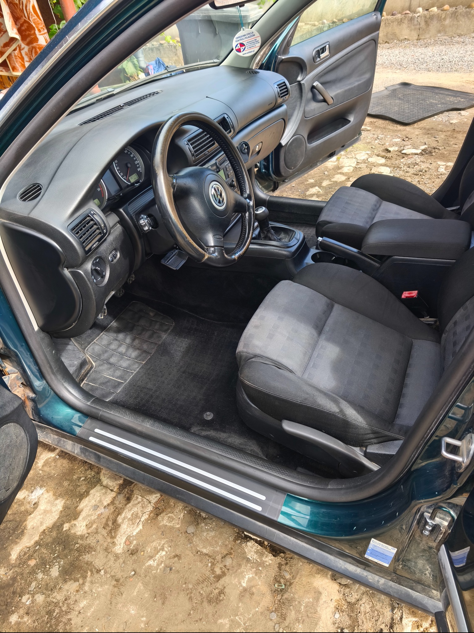 Wnętrze samochodu w kolorze butelkowej zieleni z otwartymi drzwiami. Widoczne czarne, materiałowe fotele i dywaniki. Kierownica z drewnianymi elementami.