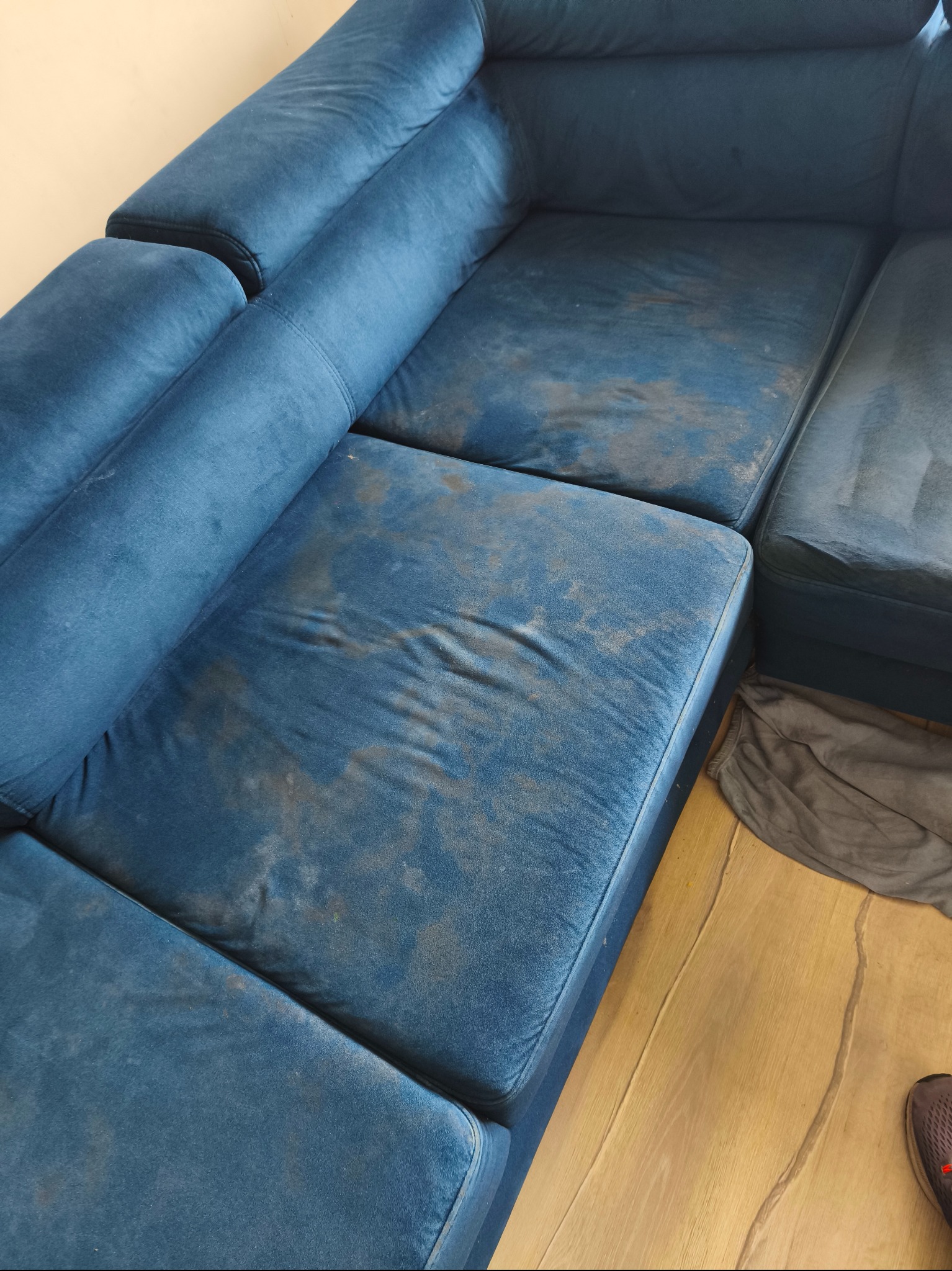 Brudna, niebieska sofa modułowa z widocznymi plamami na siedziskach, z bliska. Częściowo widoczna podłoga i szara tkanina.