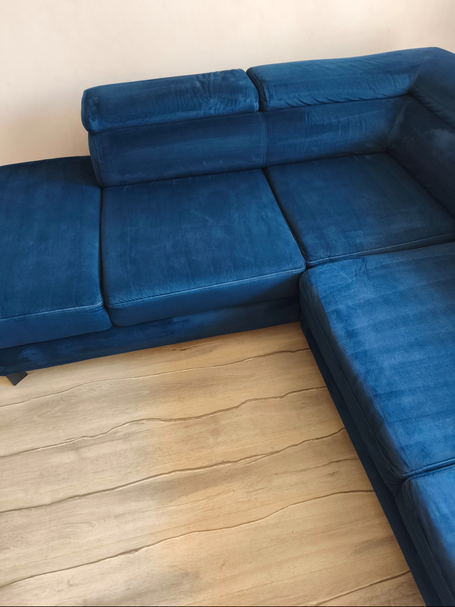 Narożnik z niebieskiego weluru na jasnej podłodze. Widoczne ślady po czyszczeniu tapicerki. Ciemniejszy odcień na siedzisku wskazuje na świeżo wyprany fragment.