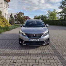 Srebrny Peugeot 5008, widok z przodu na brukowanym podjeździe, z budynkiem w tle. Tablica rejestracyjna OP 4676J. Dzień, lekkie zachmurzenie.