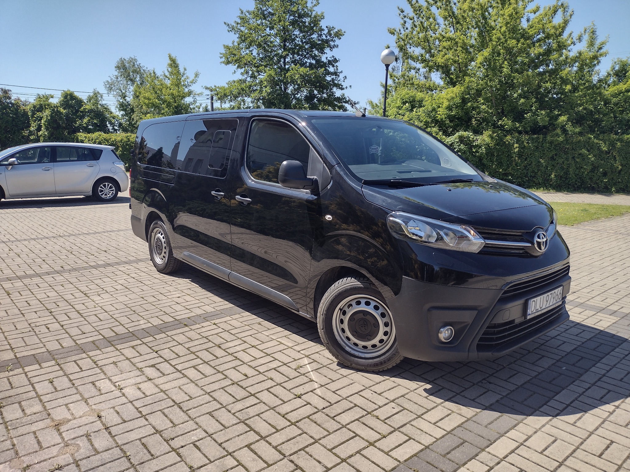 Czarny, błyszczący bus Toyota Proace Verso zaparkowany na brukowanym placu w słoneczny dzień. Widoczny fragment tablicy rejestracyjnej i zieleń w tle.