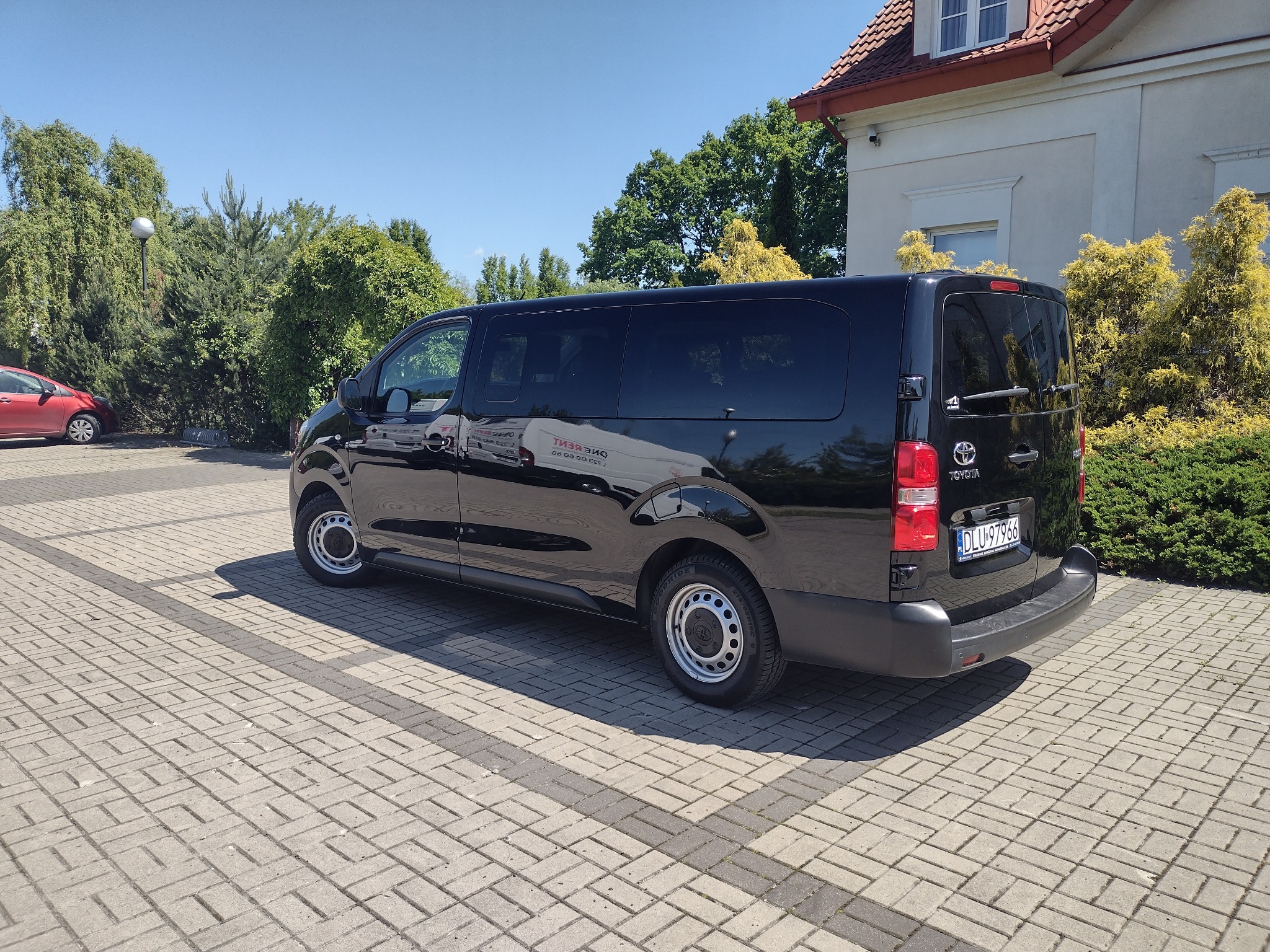 Czarny bus Toyota na kostce brukowej, zieleń w tle, tablica rejestracyjna DLU97966. Pojazd z przyciemnianymi szybami, widoczne logo Toyota.