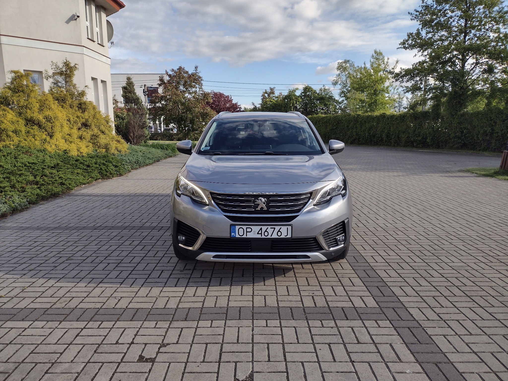 Srebrny Peugeot 5008, widok z przodu na brukowanym podjeździe, z budynkiem w tle. Tablica rejestracyjna OP 4676J. Dzień, lekkie zachmurzenie.