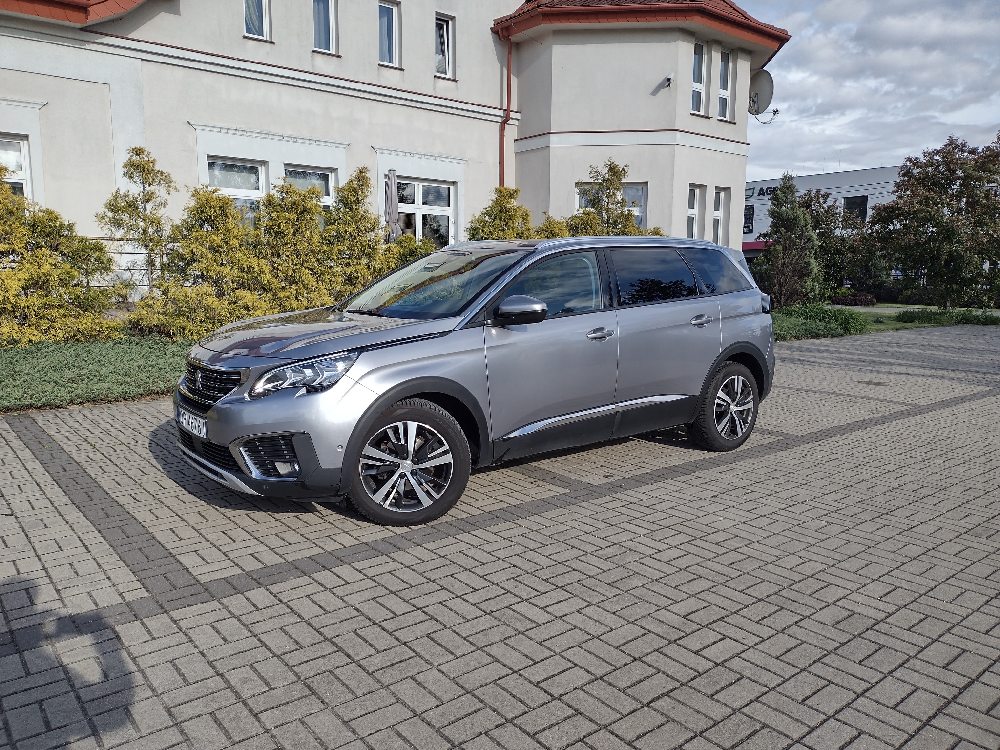 Szary Peugeot 5008 na kostce brukowej przed budynkiem z czerwonym dachem i zielenią, prezentujący przestronność i elegancję transportu rodzinnego.