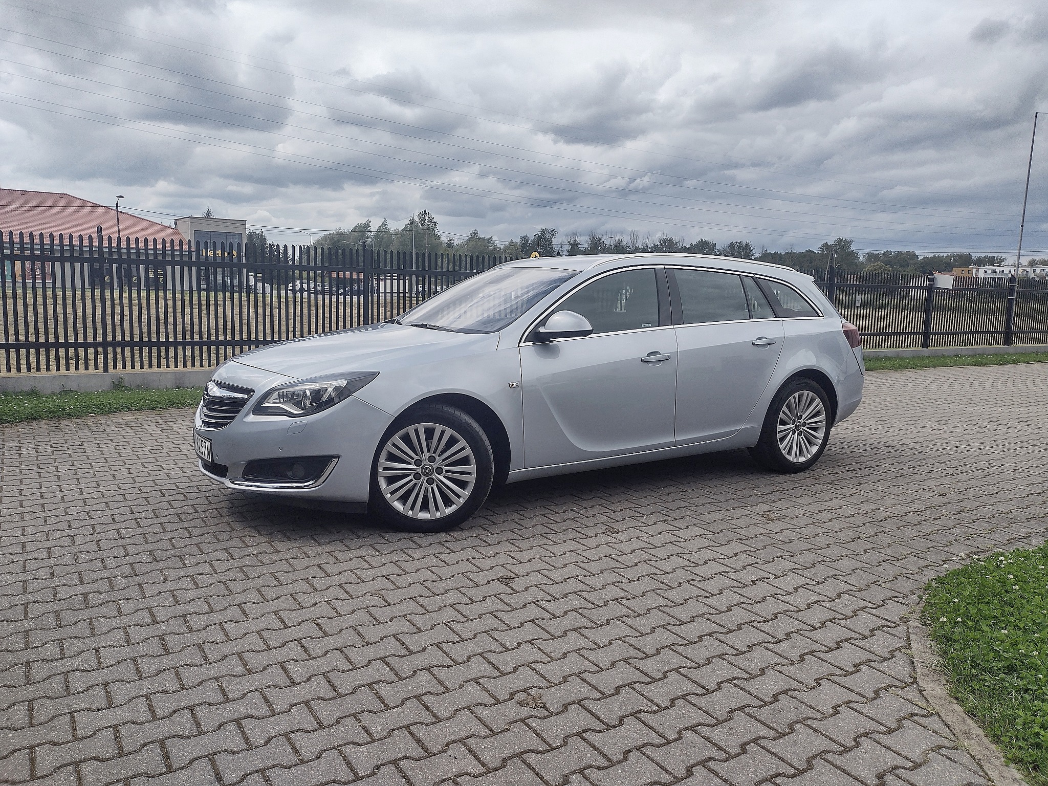Srebrna Opel Insignia kombi na kostce brukowej, z widocznym ogrodzeniem i pochmurnym niebem w tle. Auto z relingami dachowymi i aluminiowymi felgami.