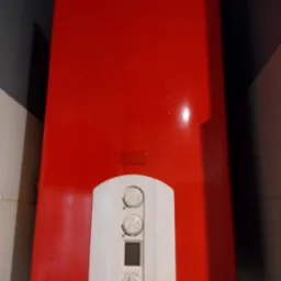 Czerwony piec gazowy z białym panelem sterowania na tle szarej ściany i białych płytek. Widoczny fragment rury odprowadzającej spaliny.