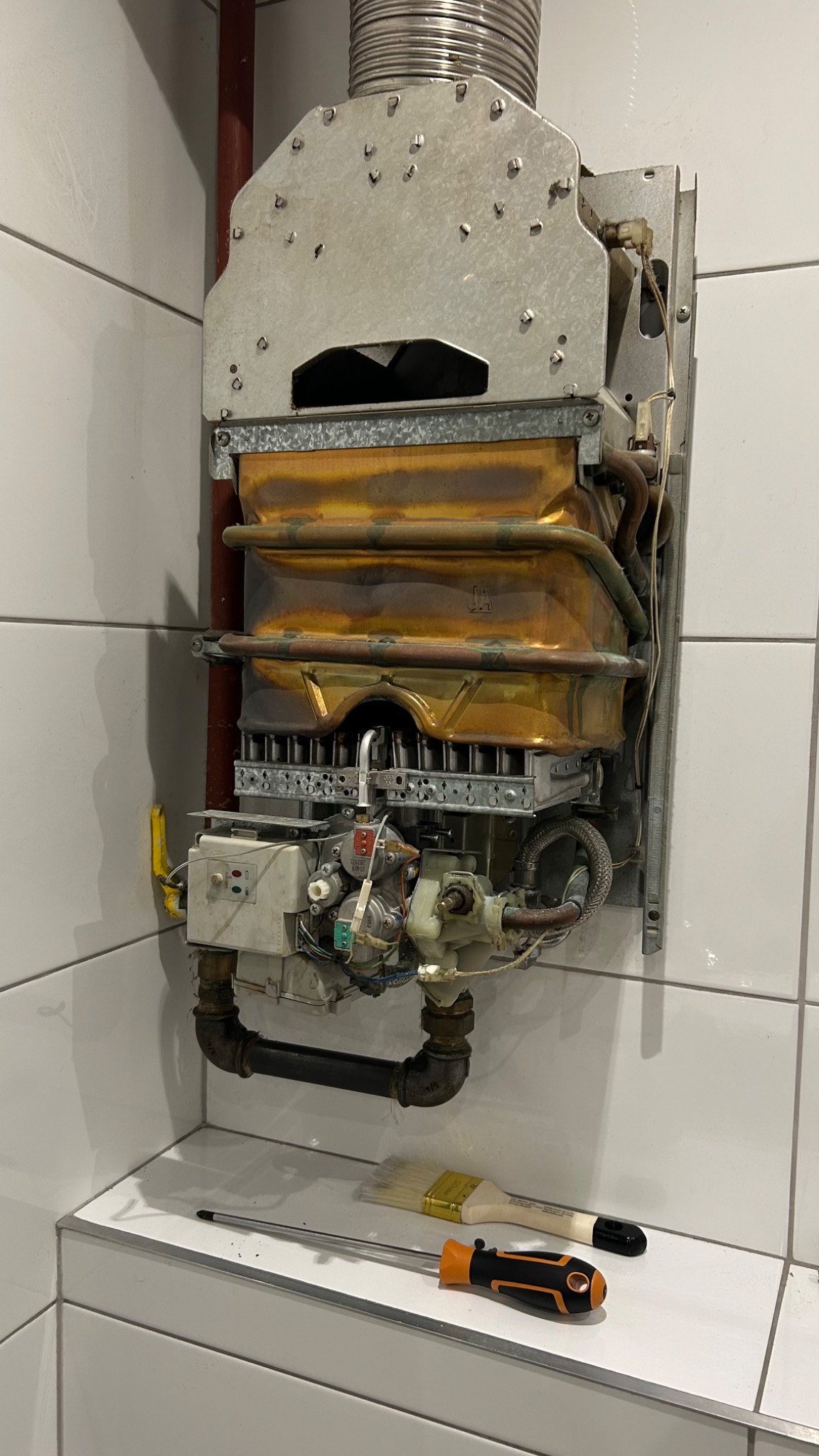 Otwarty piec gazowy z widocznymi elementami wewnętrznymi, narzędzia leżą na półce. Widoczne ślady użytkowania na elementach pieca.