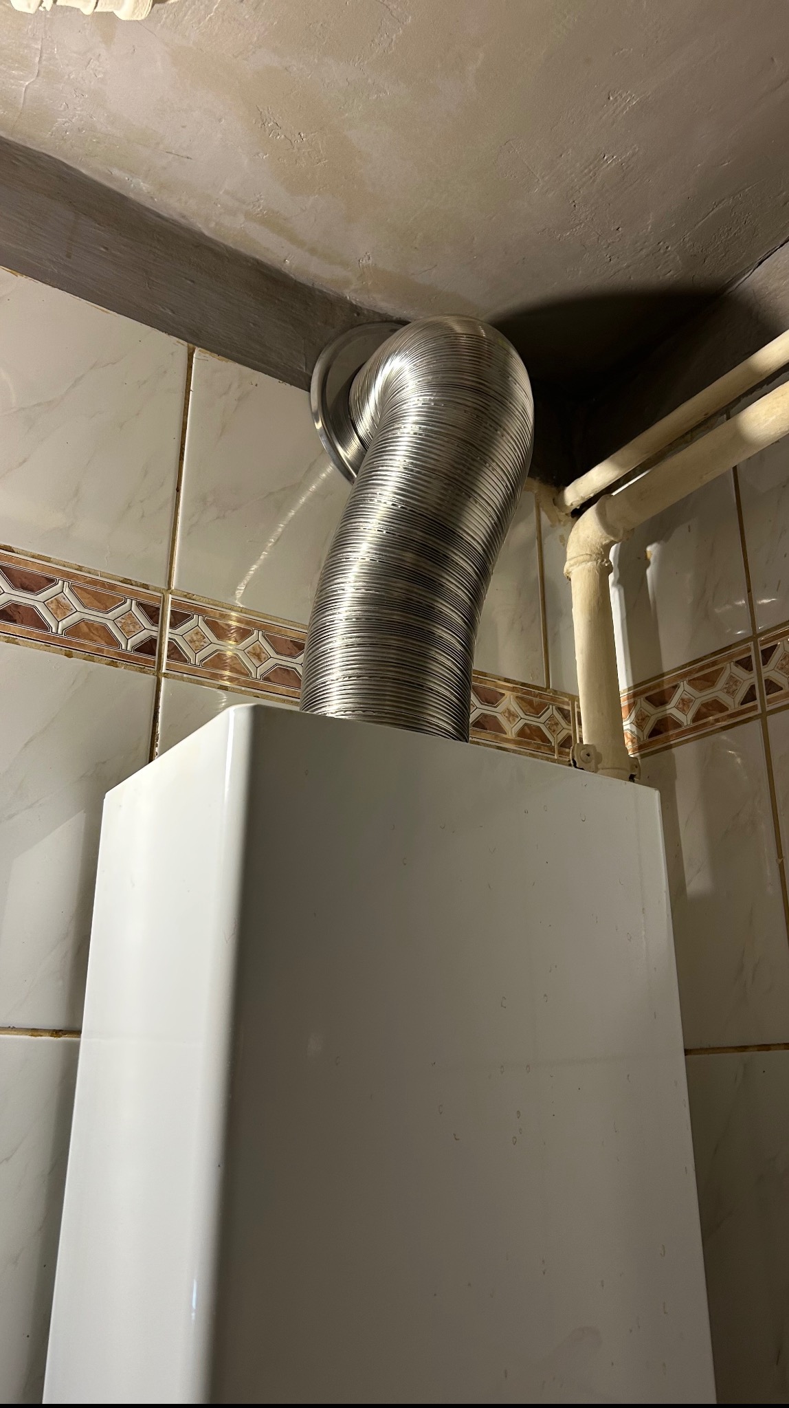 Biały piec gazowy z podłączonym, elastycznym, metalowym przewodem spalinowym, zamontowany w pomieszczeniu z kafelkami. Widoczne rury instalacyjne.