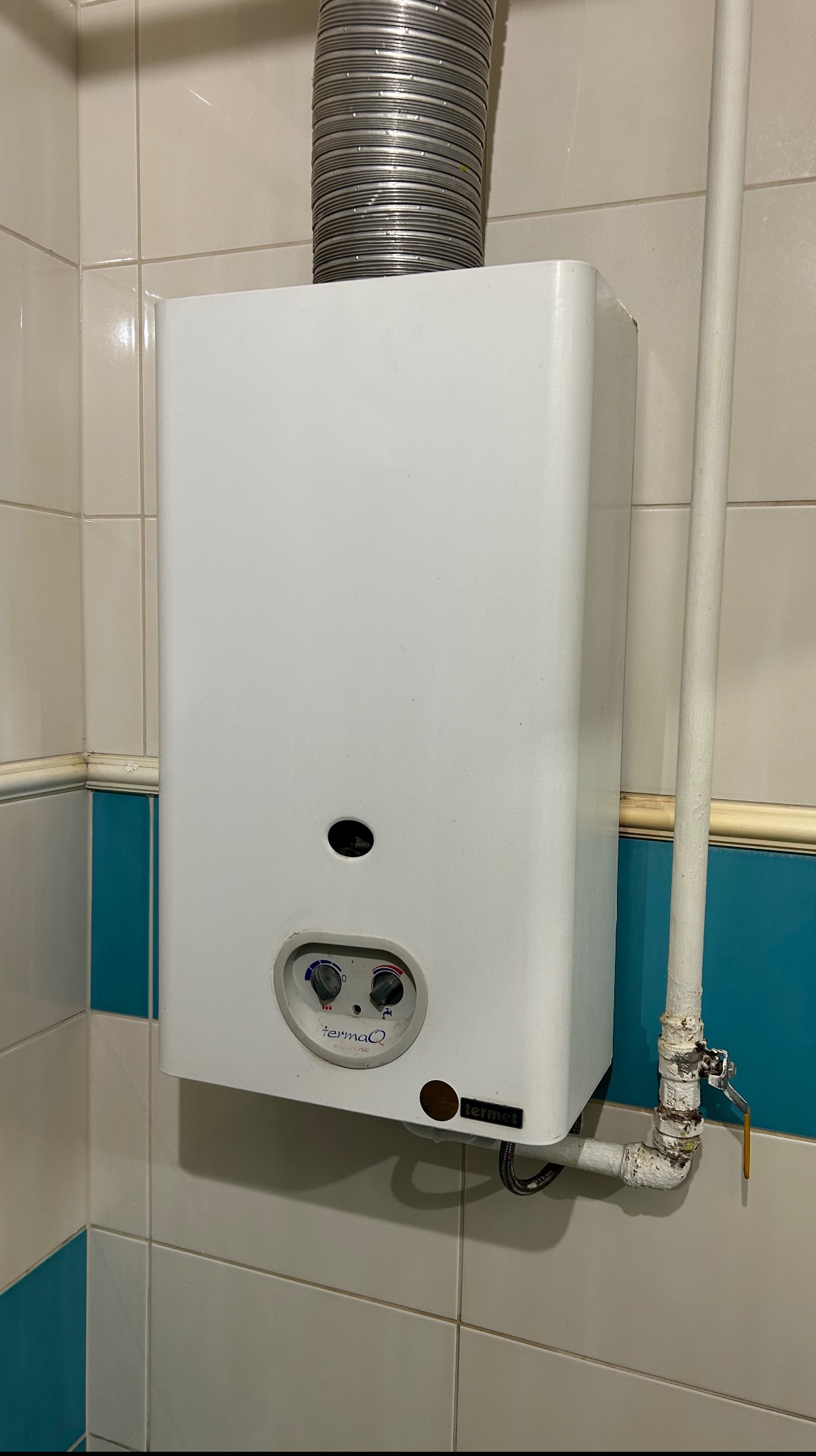 Biały piec gazowy TermaQ z szarą rurą odprowadzającą spaliny, zamontowany na kafelkowanej ścianie z niebieskim akcentem. Widoczne przyłącza gazowe i wodne.