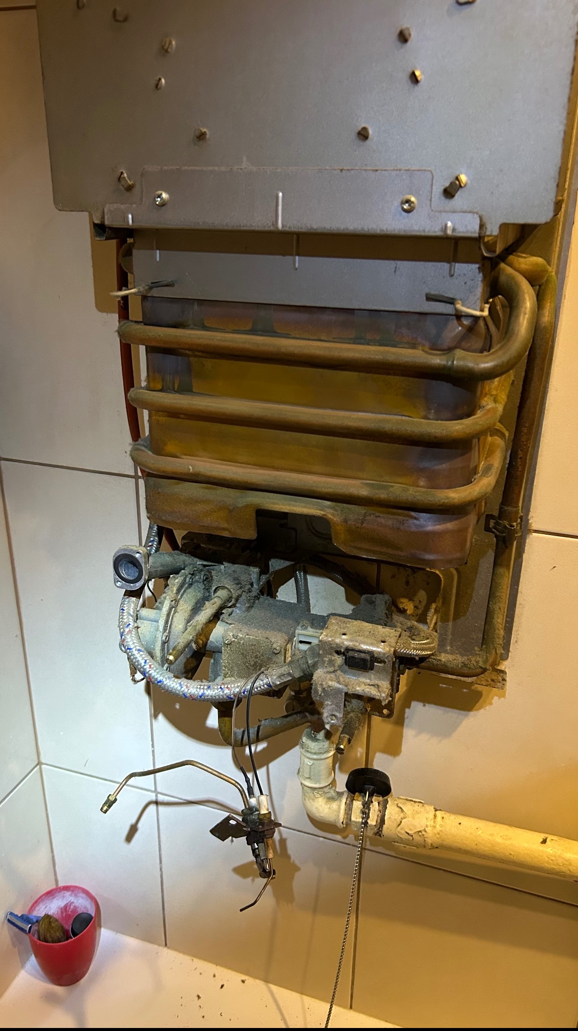 Widok na zdemontowany, stary piec gazowy z widocznymi elementami instalacji i przewodami, sugerujący etap serwisu lub naprawy. Białe kafelki w tle.