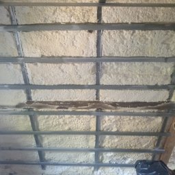 Krzysztof Gardzielik - Izolacja poddasza pianą PUR między metalowymi profilami konstrukcyjnymi. Widoczna struktura piany i fragment muru. Ocieplenie stropu pianką poliuretanową.