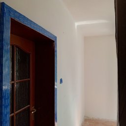 Krzysztof Gardzielik - Przygotowanie do malowania: drewniane drzwi z szybą zabezpieczone niebieską taśmą malarską, ściana świeżo pomalowana na biało, widoczne drobne niedoskonałości.